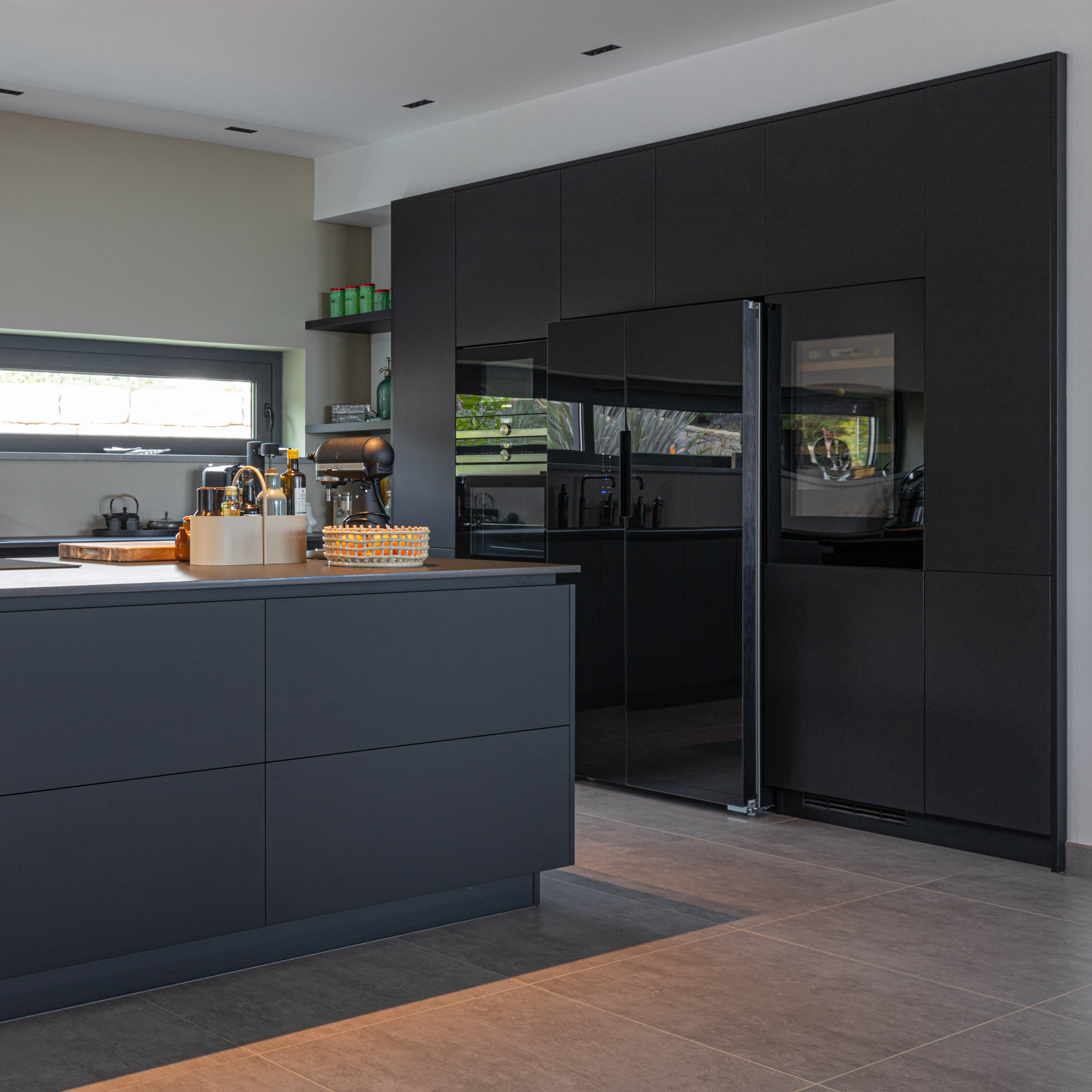 Cozinha com garrafeira e americano em vidro preto Cozinha JMQ luxuosa no Algarve. Cozinha com frentes em Fenix preto, bancada em Neolith, sistema BORA Pure, garrafeira Liebherr e equipamentos Siemens.